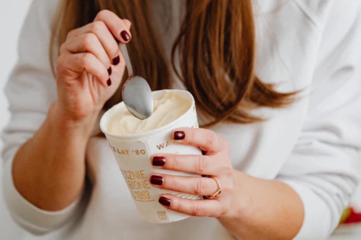 Food cravings. A woman with long brown hair, wearing a light top, holding a tub of ice cream in one hand and scooping it with a spoon in the other. 