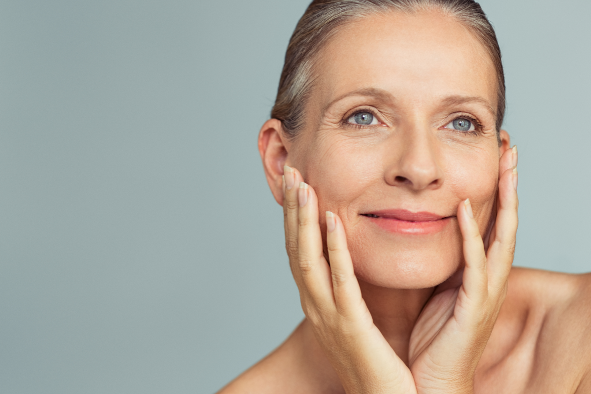 Confident midlife woman with glowing, healthy skin gently touching her face and smiling. Represents natural skincare, graceful ageing, and inner vitality.