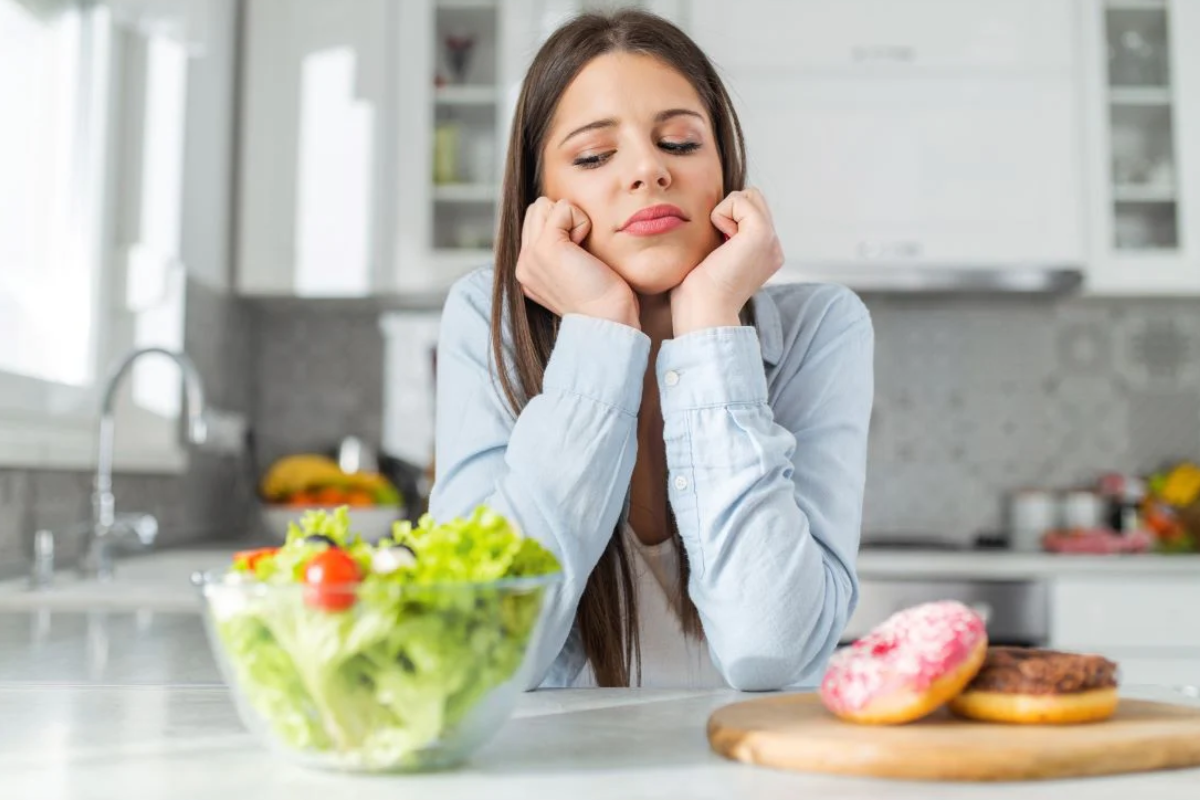 Food cravings. Young woman sitting at a kitchen bench with her chin resting on her hands, looking frustrated while choosing between a bowl of fresh salad and a plate of donuts.